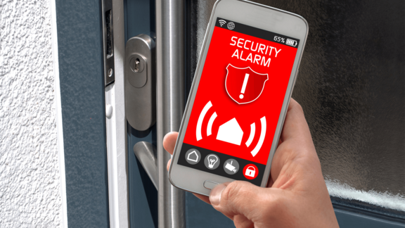 Alarmanlage per App steuern – volle Kontrolle über Ihr Zuhause. Smartphone mit geöffneter Sicherheits-App vor Hauseingangstür, Symbol für moderne Alarmanlagensteuerung per App.